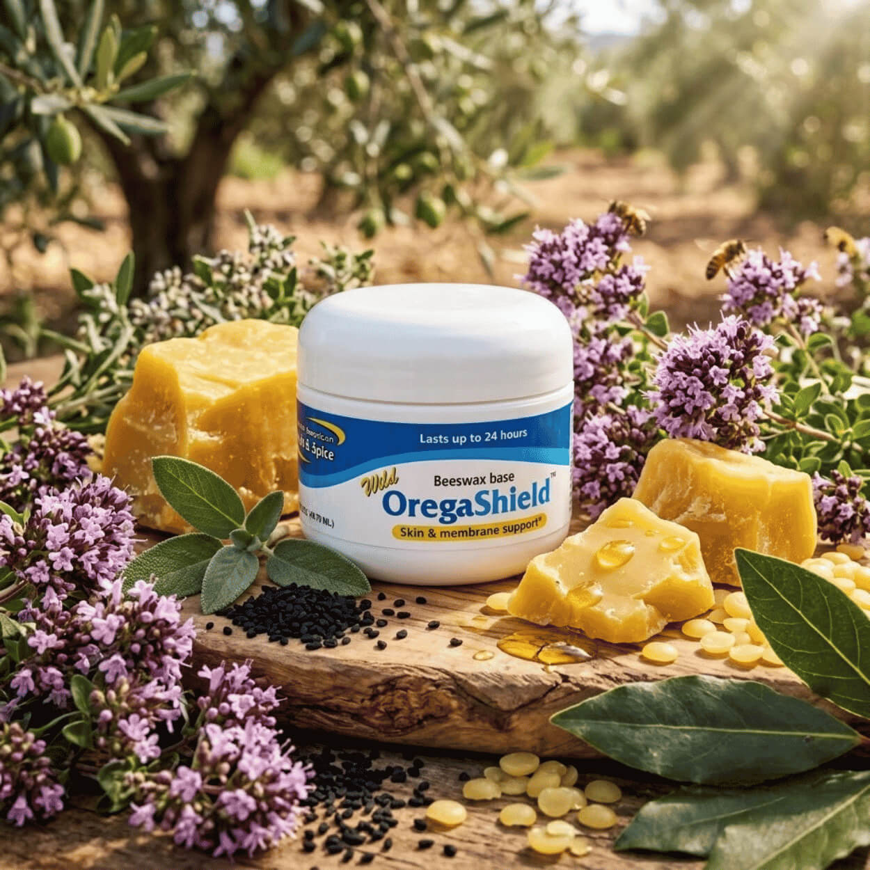 A white jar of OregaShield balm stands on a wooden surface surrounded by pieces of beeswax, purple oregano flowers with flying bees, leaves, and black seeds with an olive grove in the background.